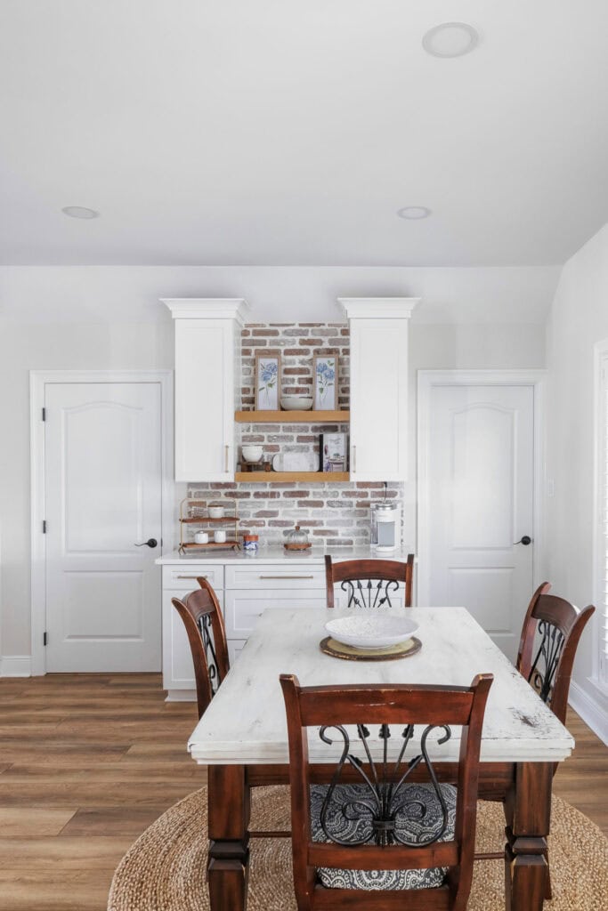 Rustic kitchen nook with white custom cabinets, brick backsplash, open shelving, and a farmhouse-style dining table.