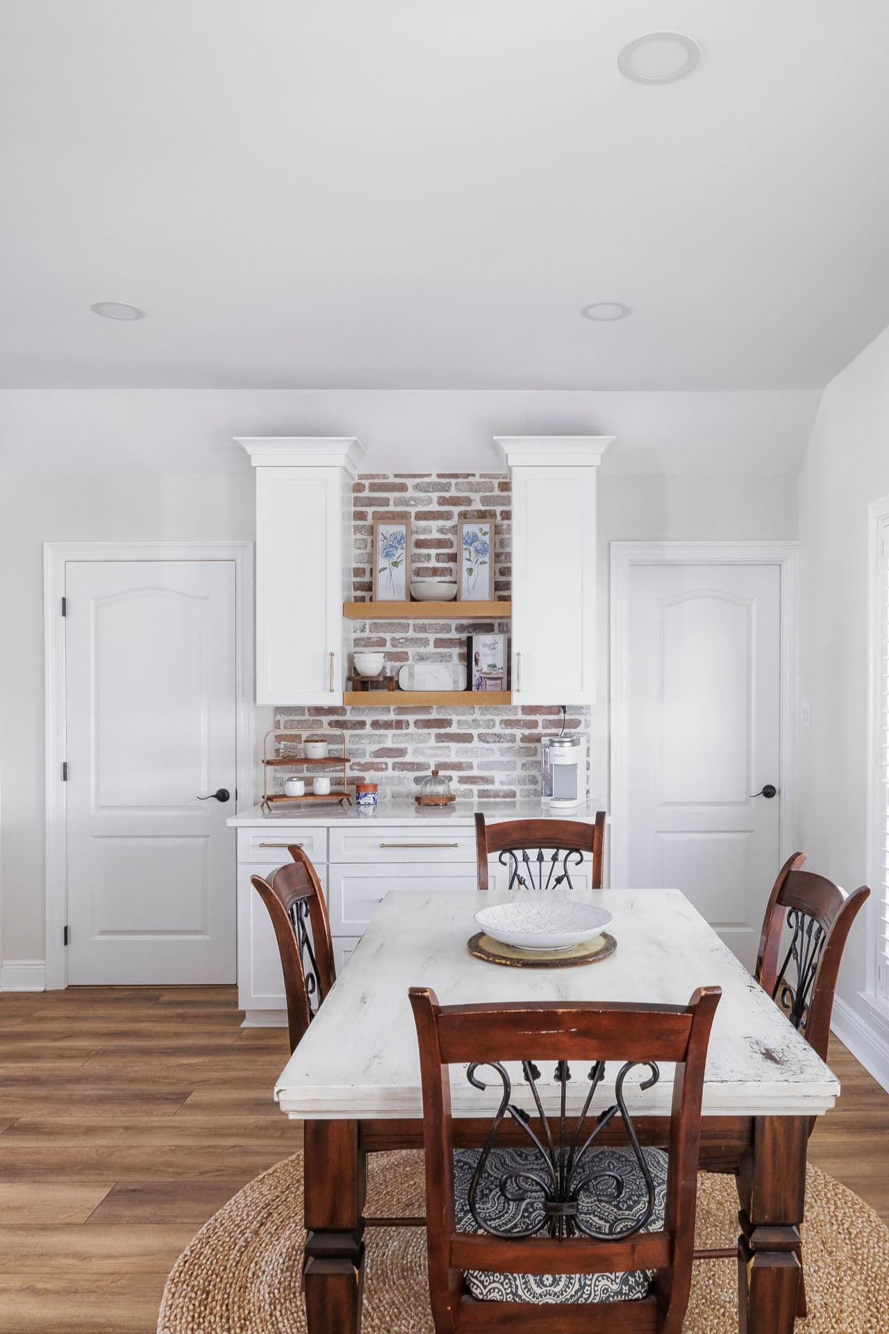 Rustic kitchen nook with white custom cabinets, brick backsplash, open shelving, and a farmhouse-style dining table.