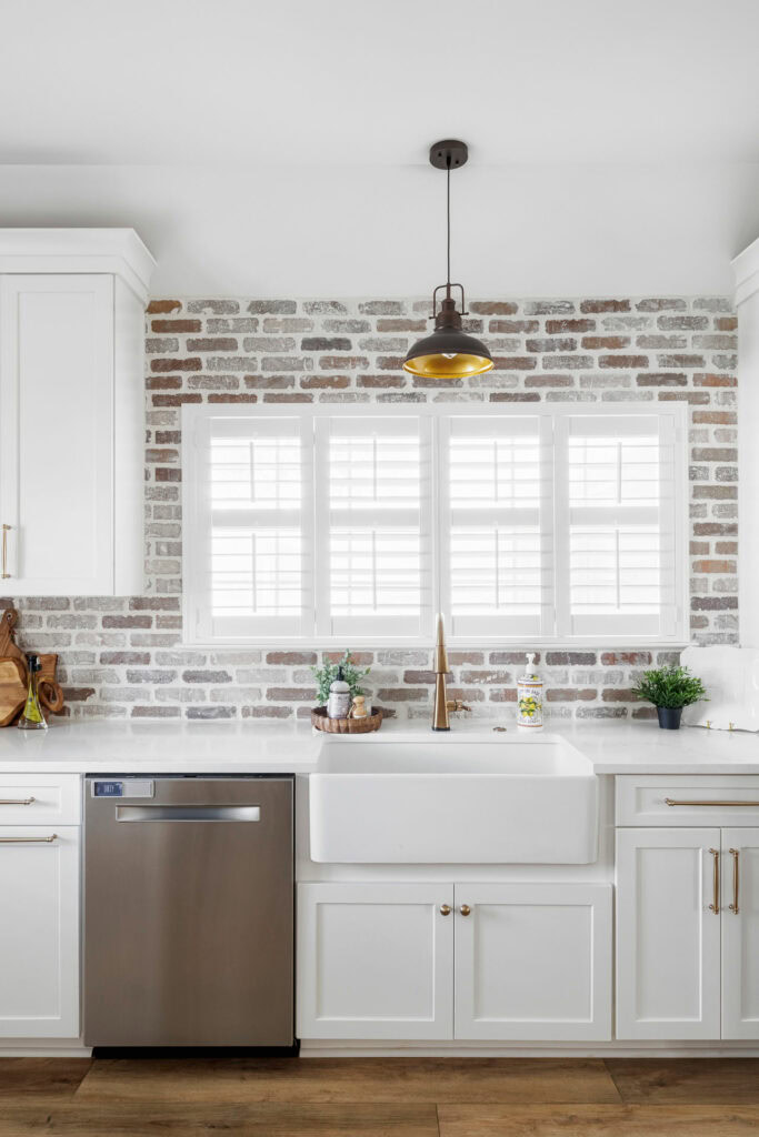 Close-up of white farmhouse sink with brass faucet in a modern kitchen featuring white shaker cabinets, stainless steel dishwasher, and a rustic brick backsplash