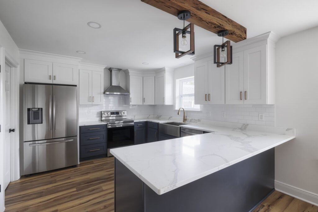 Modern two-tone kitchen featuring navy and white shaker cabinets, marble-look quartz countertops, a stainless steel farmhouse sink, and a rustic wood beam with pendant lights.