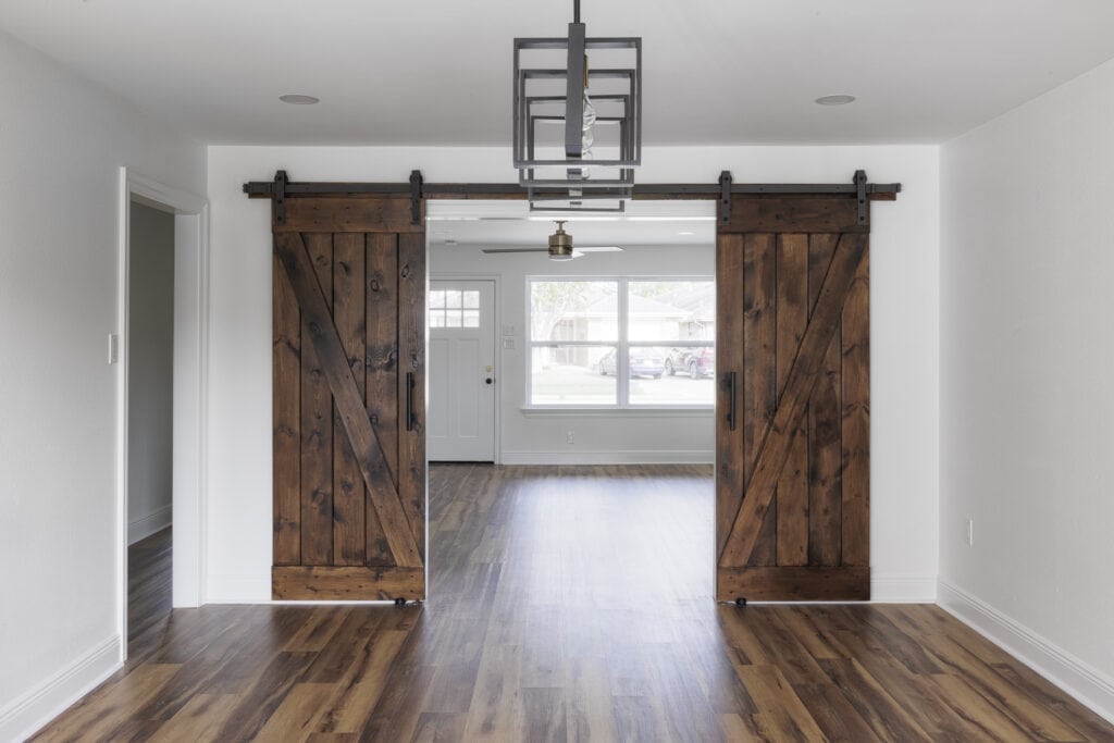 Double rustic barn doors on black metal tracks open to a bright living area with natural light, wood flooring, and a modern square-frame pendant light.
