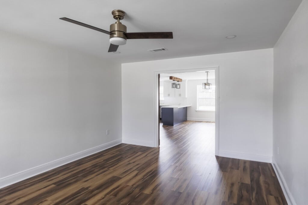 Transitional room with wood-look flooring, modern ceiling fan, and a view into a bright kitchen with exposed wood beam and navy island.