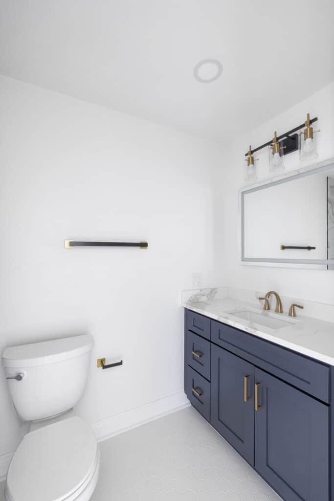 Toilet and navy vanity with marble countertop, gold fixtures, and LED mirror in a bright white bathroom.