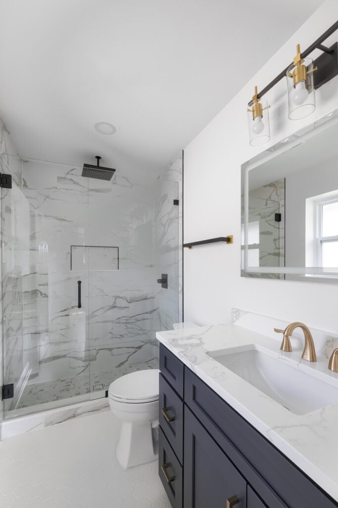 Modern bathroom remodel with walk-in shower, black rain showerhead, frameless glass enclosure, navy vanity with marble countertop, and gold fixtures.
