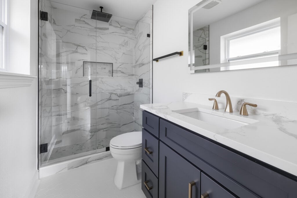 Modern bathroom with marble-look tile shower, navy vanity, and brushed gold fixtures.