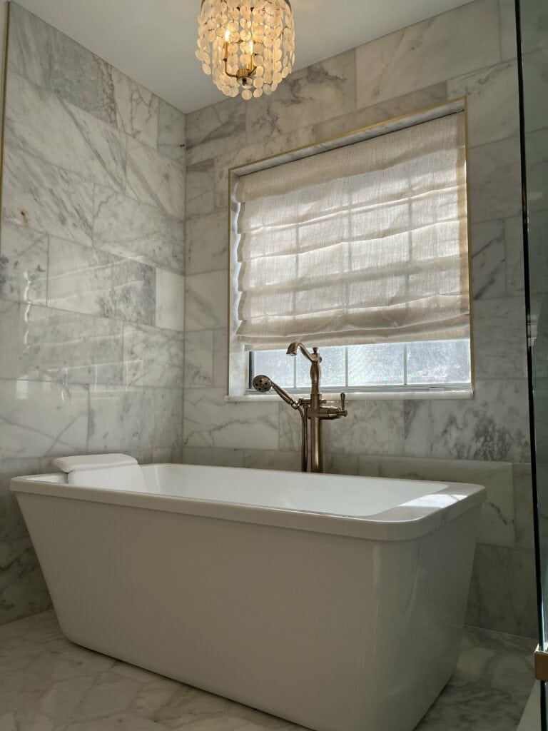 Freestanding tub beneath chandelier with marble walls and roman shade