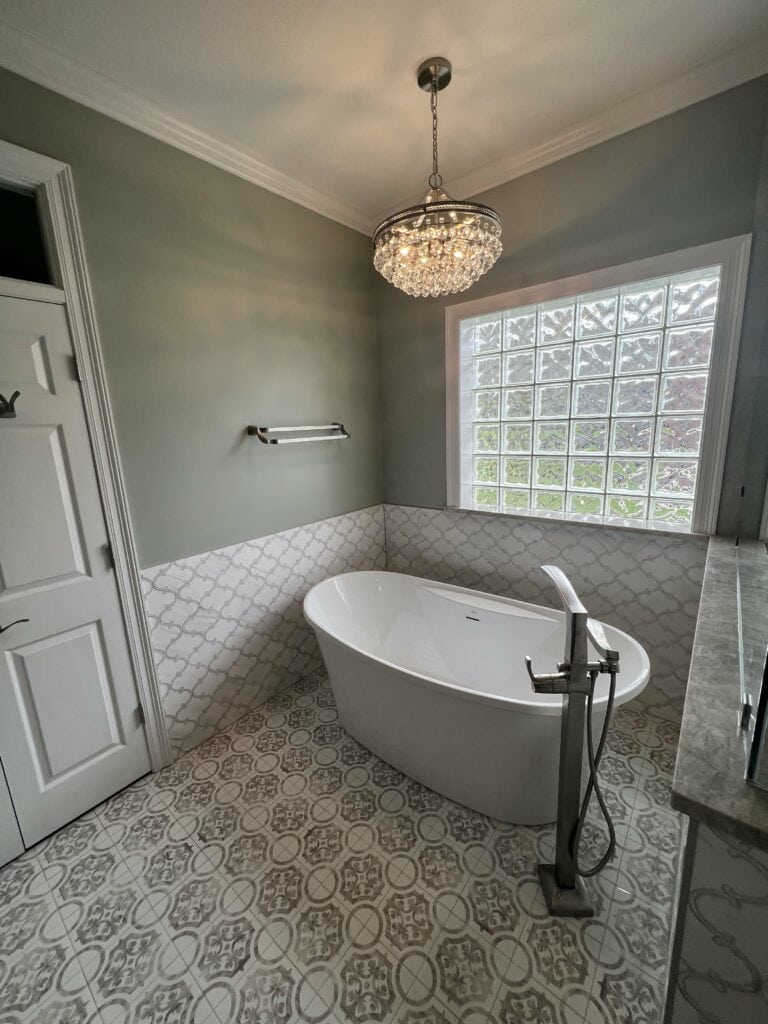 Freestanding white soaking tub beneath a crystal chandelier with glass block window and decorative tile surround.