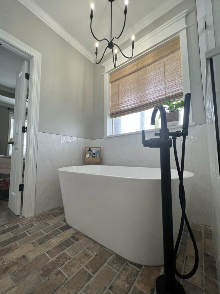 Freestanding soaking tub with matte black floor-mounted faucet, brick flooring, and hexagon wall tile accent.