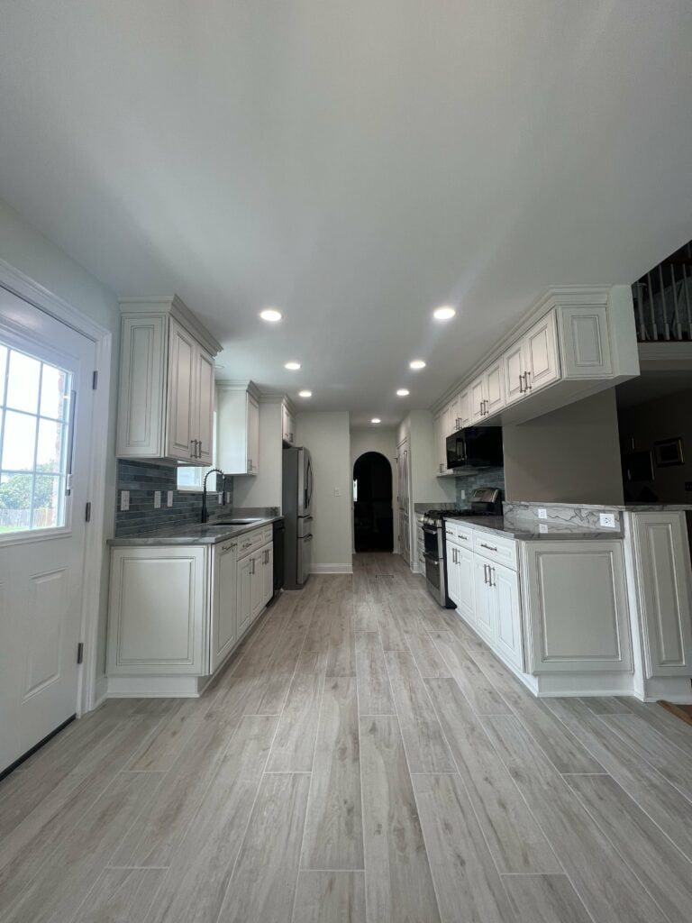 Wide-angle view of a remodeled kitchen with custom white cabinetry, wood-look tile flooring, and stainless steel appliances.