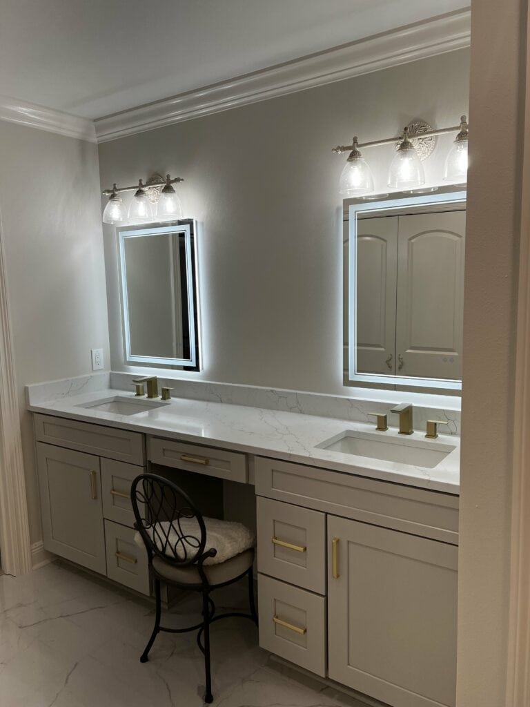 Double vanity with gold hardware and backlit mirrors in luxury bathroom.
