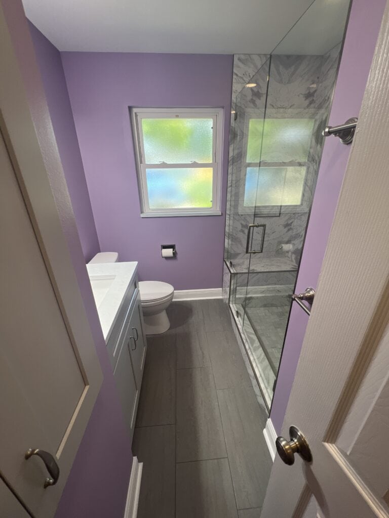 Compact bathroom remodel with glass walk-in shower, gray tile flooring, and lavender walls.