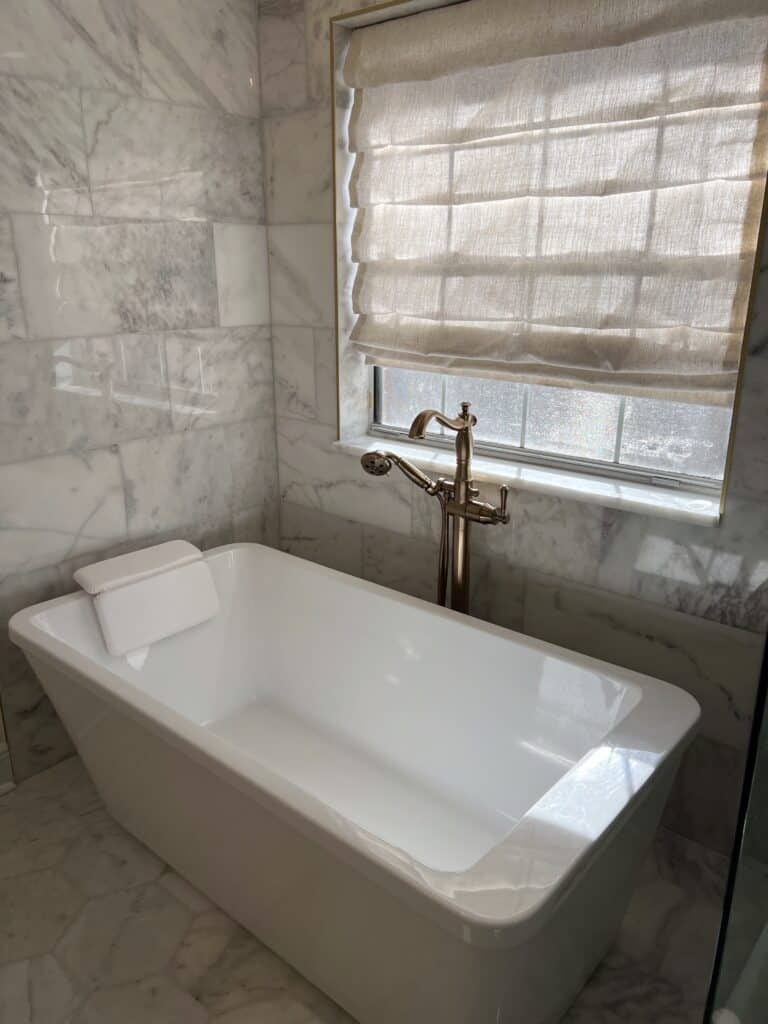 Freestanding soaking tub with brass filler in a marble-tiled bathroom.
